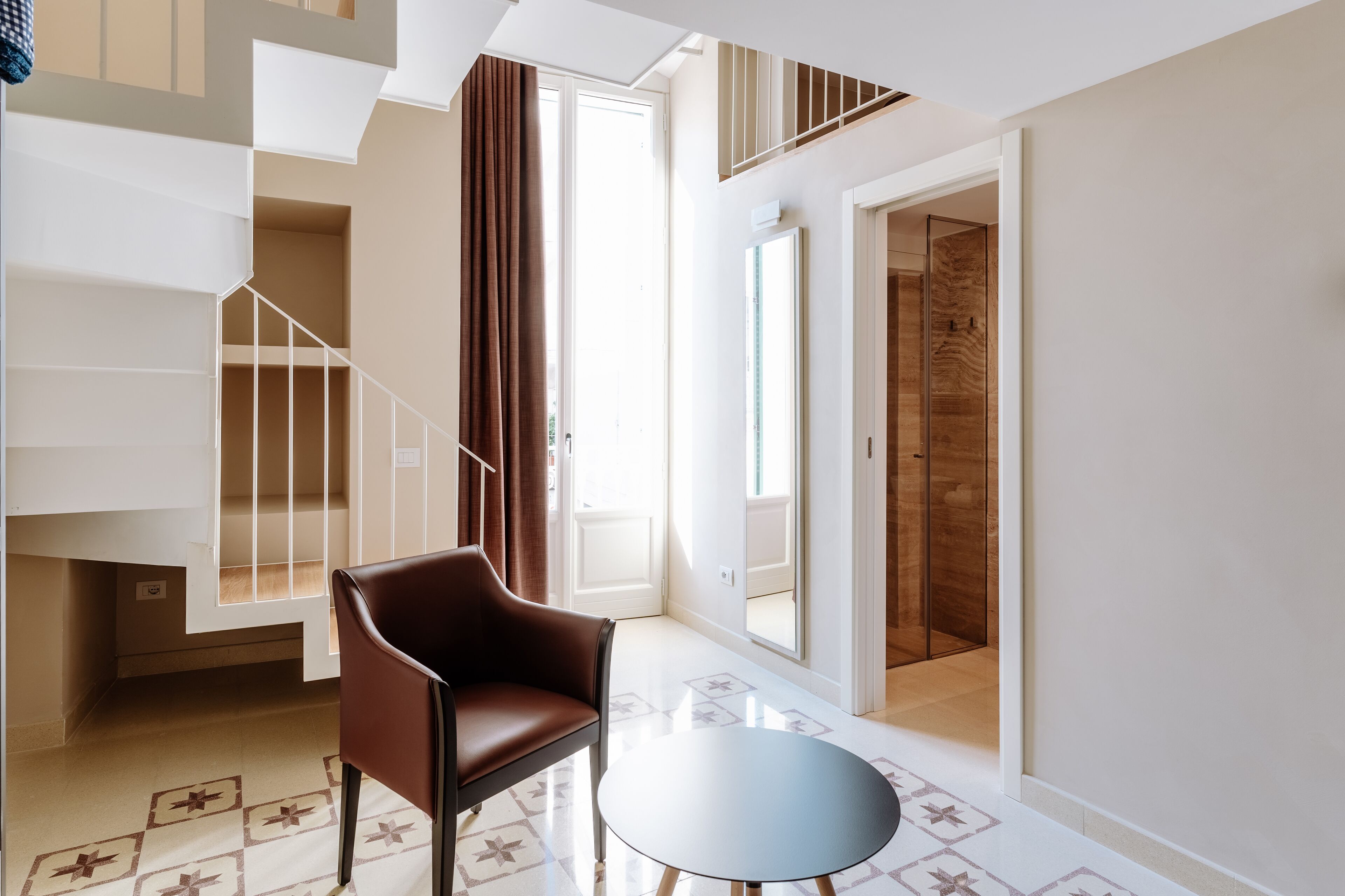 Une chambre d’hôtel moderne avec un escalier, un fauteuil en cuir marron, une petite table ronde et un sol à motifs.