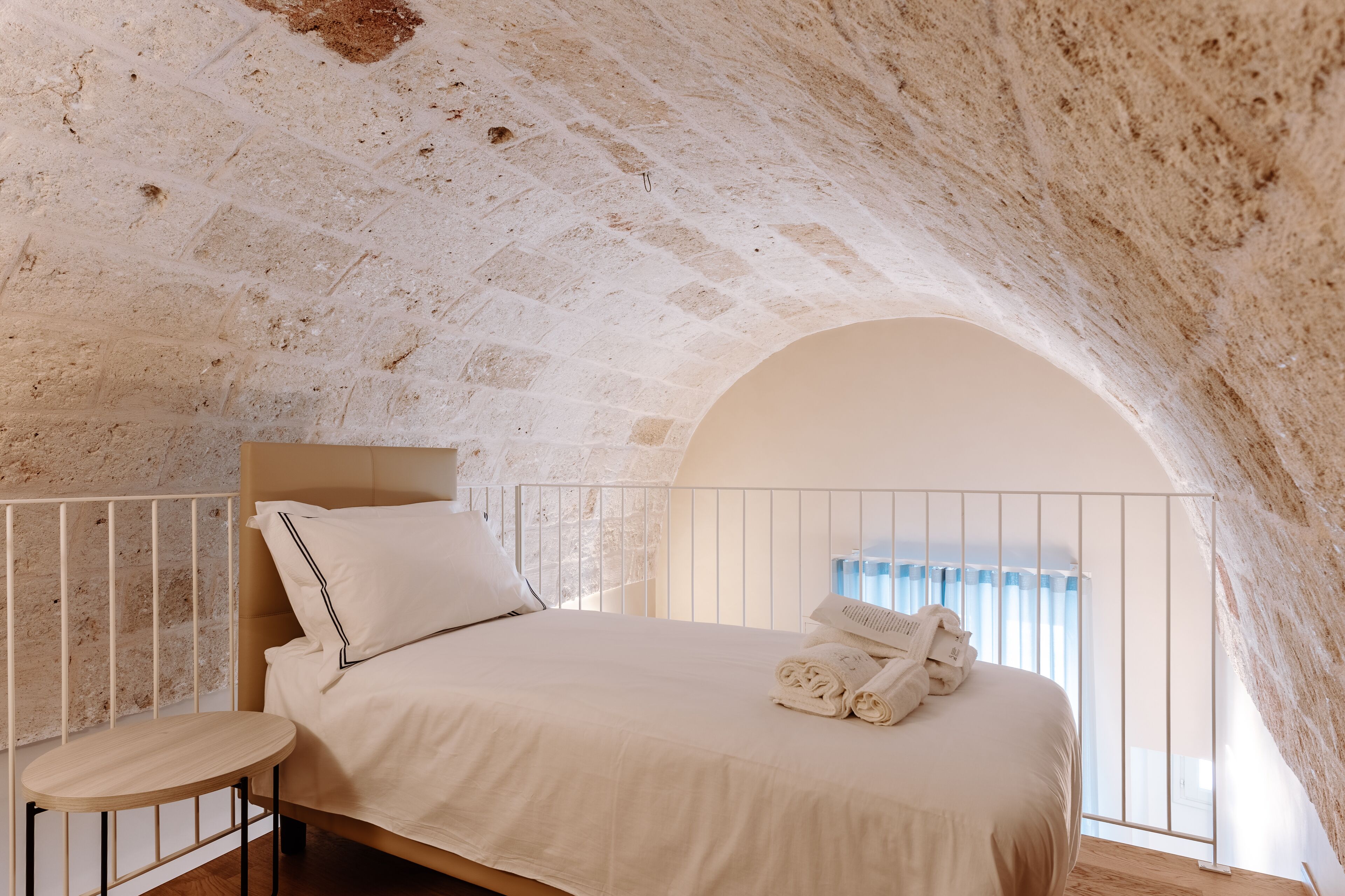 A bedroom with a stone ceiling, a bed with white linens, a small round table, and a view of the sky through an arched window.