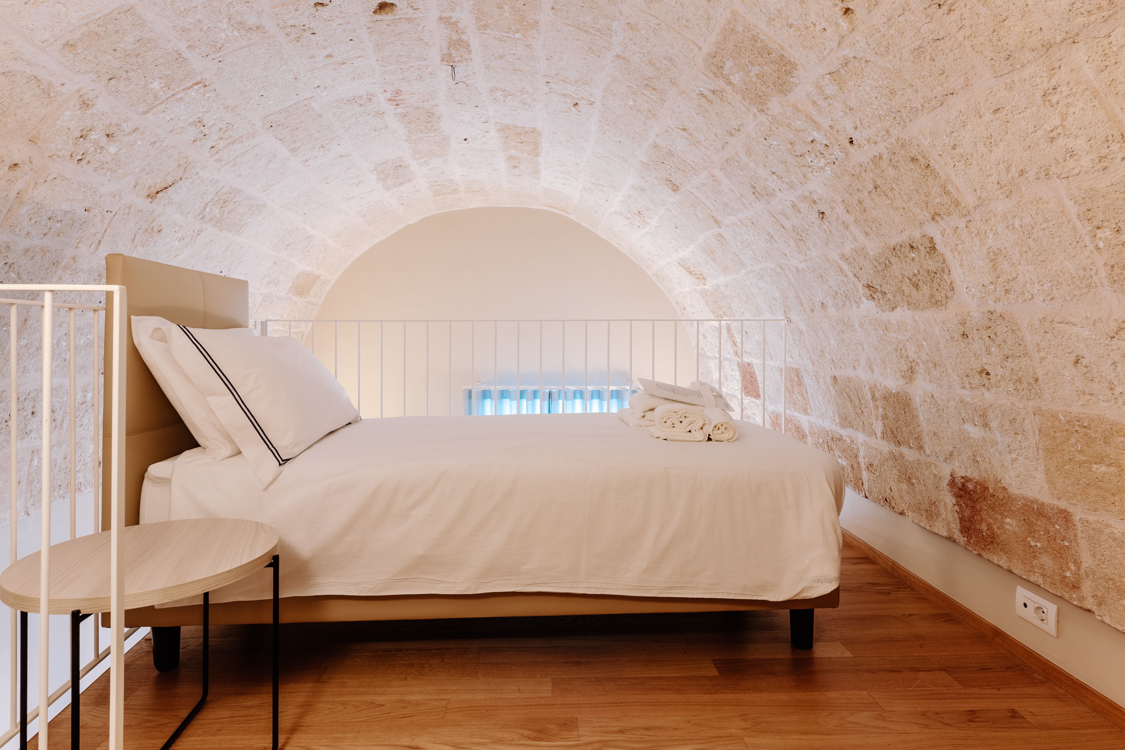 A bedroom with a stone arch ceiling, a bed with white bedding, a bedside table, and a window with blinds.