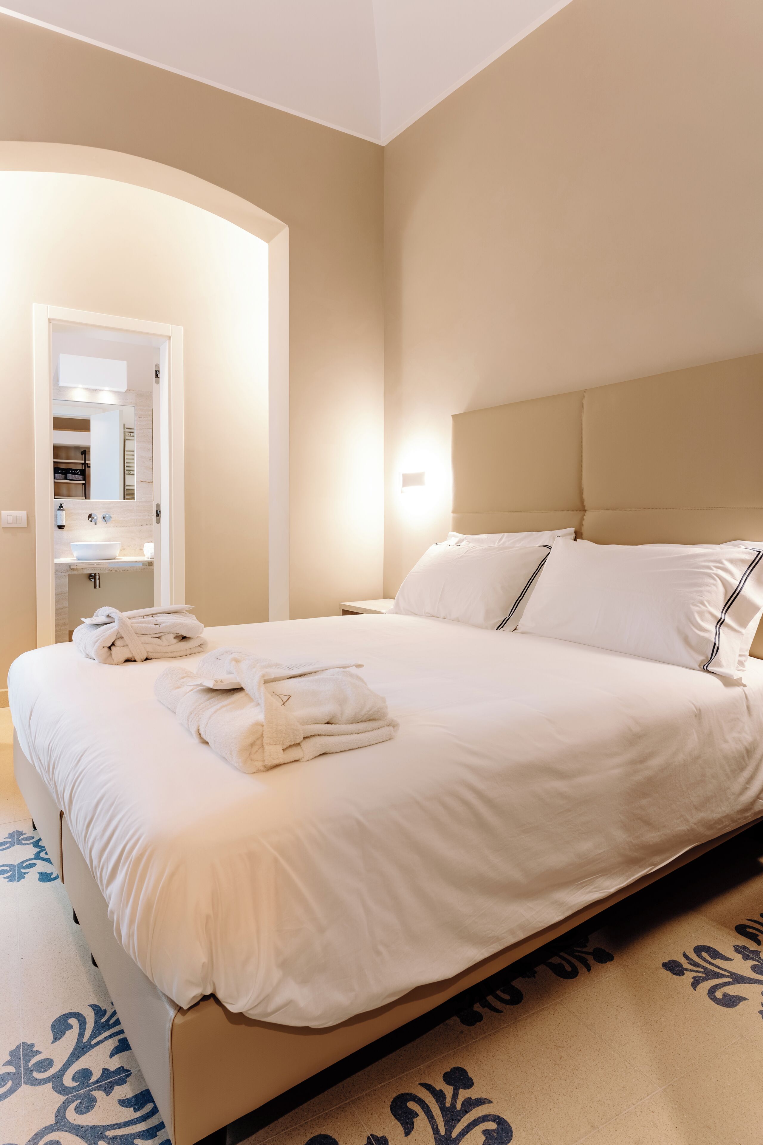 A neatly made bed with white linens and a headboard, a bathroom with a sink and mirror, and a patterned rug on the floor.