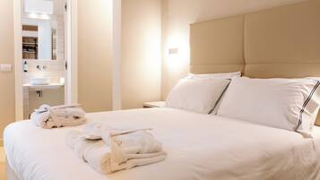 A neatly made bed with white linens and a headboard, a bathroom with a sink and mirror, and a patterned rug on the floor.