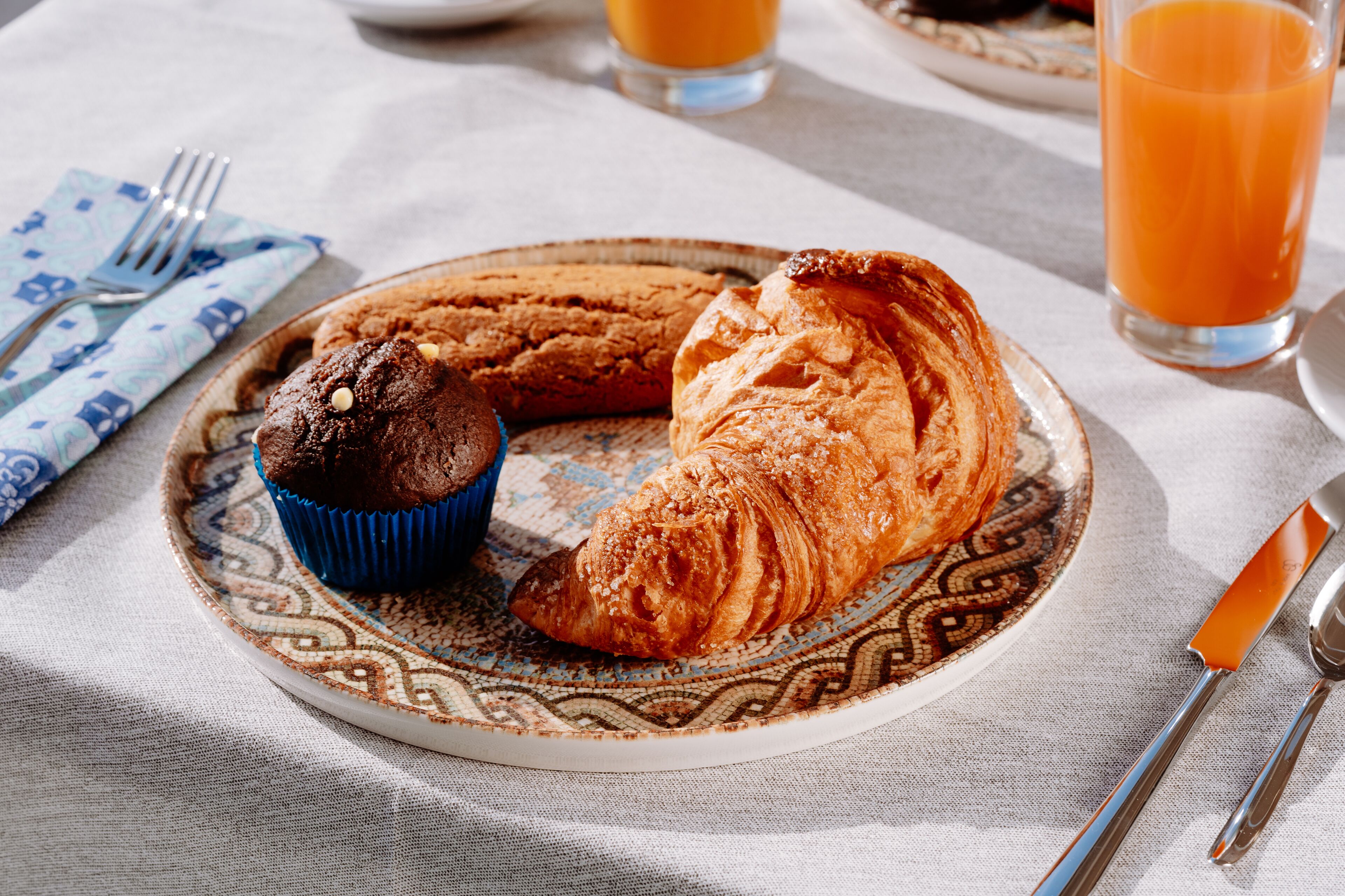 Une assiette avec un croissant et un muffin, posée sur une nappe à motifs, un verre de jus d’orange et des couverts sur une table.