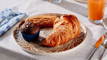 Une assiette avec un croissant et un muffin, posée sur une nappe à motifs, un verre de jus d’orange et des couverts sur une table.