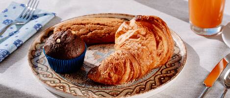 Une assiette avec un croissant et un muffin, posée sur une nappe à motifs, un verre de jus d’orange et des couverts sur une table.