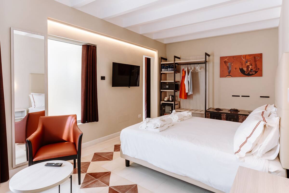 A hotel room with a large bed, a seating area with a chair, a television, a wardrobe, and a painting on the wall.