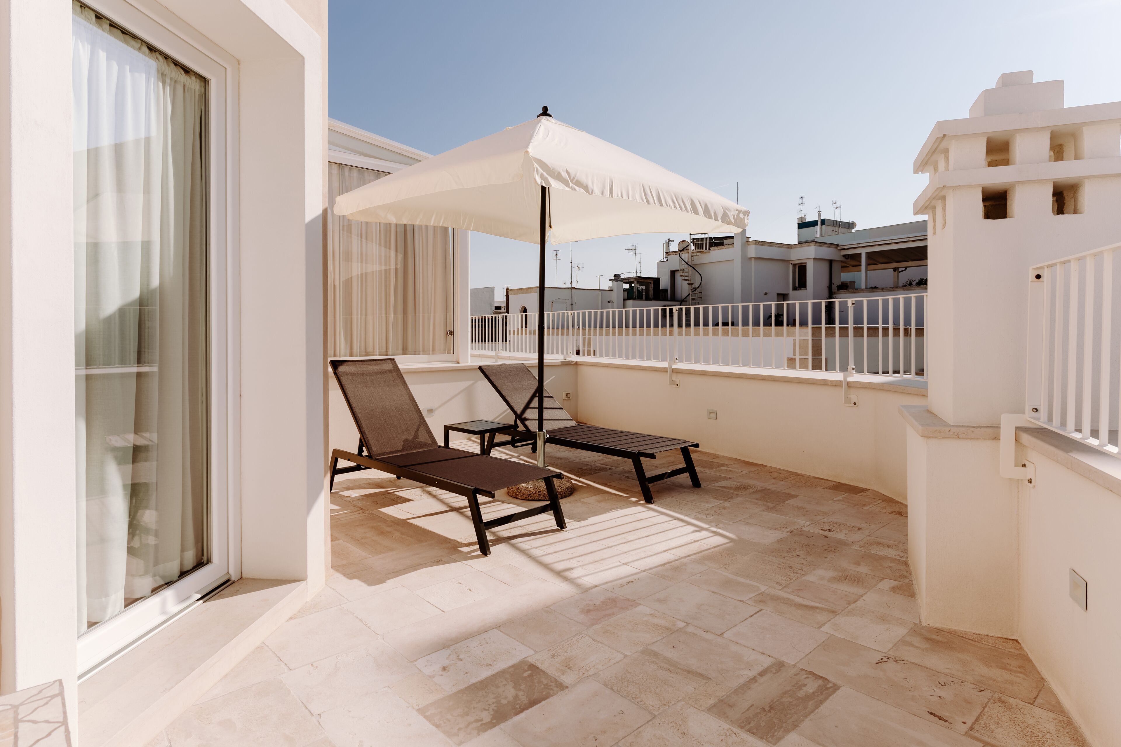 A balcony with two sun loungers and a white umbrella, a tiled floor, and a view of neighboring buildings.