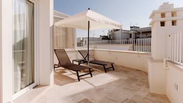 A balcony with two sun loungers and a white umbrella, a tiled floor, and a view of neighboring buildings.