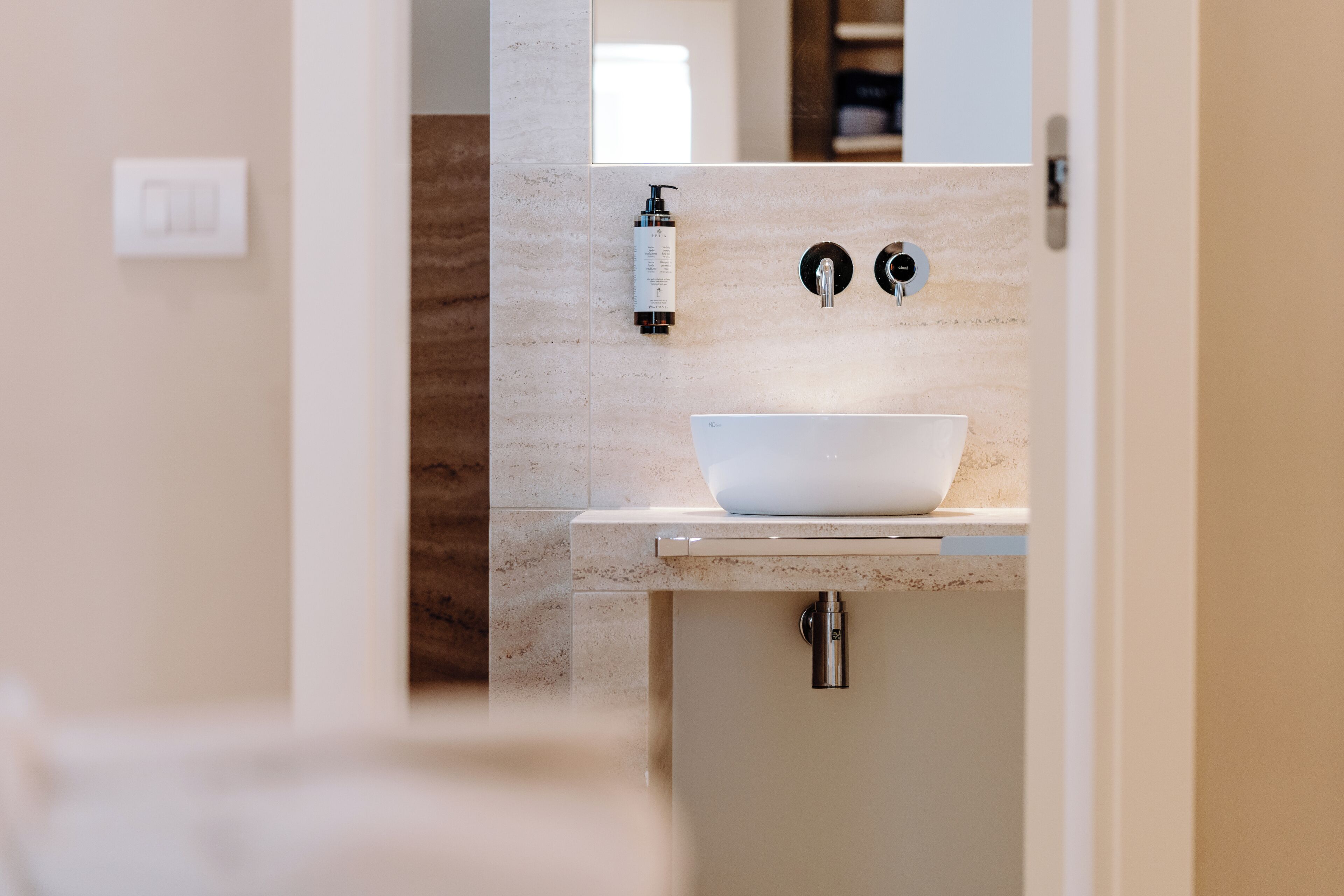 A modern bathroom with a large white basin, a handheld showerhead, and a bottle of shampoo on the wall.