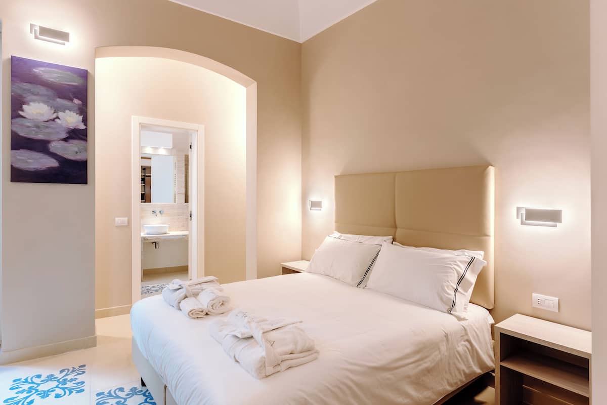 A neatly made bed with white linens and towels, a headboard, bedside tables, and a bathroom visible through an archway.