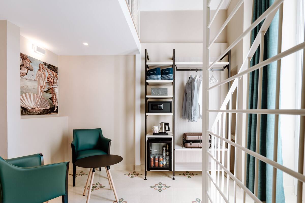 A modern hotel room with a green sofa, a small table, a minibar, and a painting on the wall.