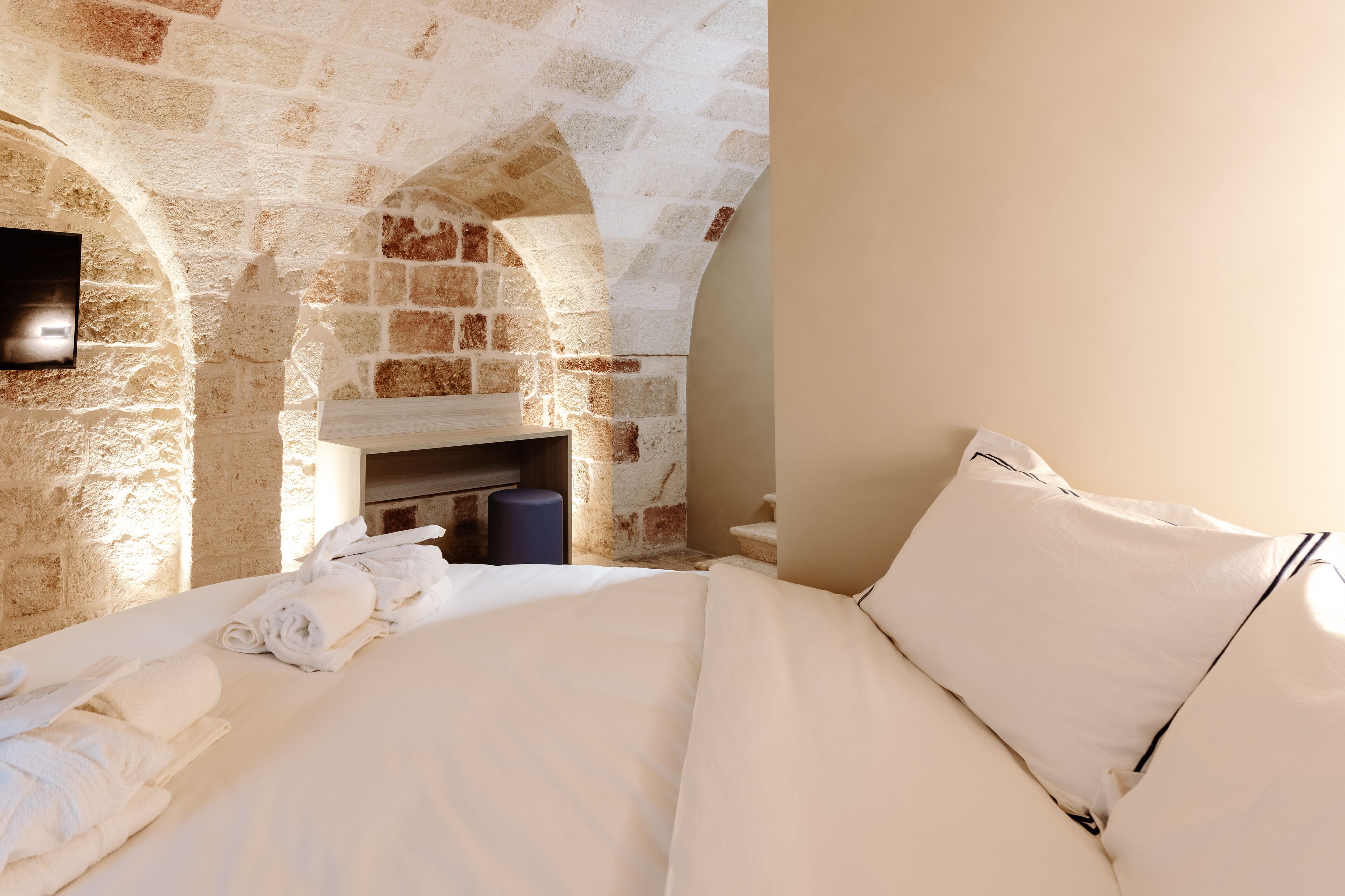Una habitación con pared de piedra, chimenea, cama con sábanas blancas y un objeto cilíndrico azul en el suelo.