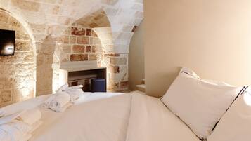 Una habitación con pared de piedra, chimenea, cama con sábanas blancas y un objeto cilíndrico azul en el suelo.