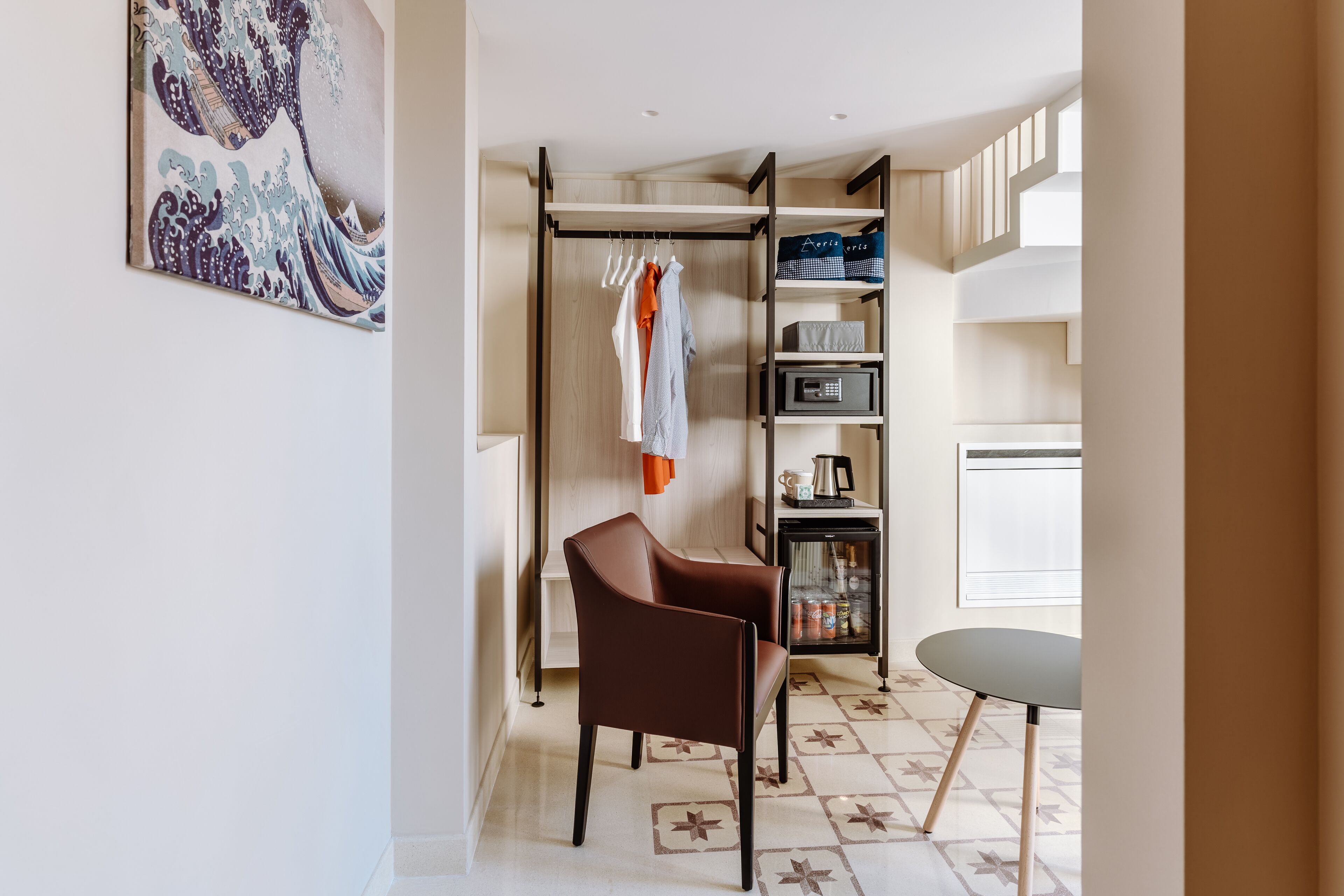 A modern room with a brown leather chair, a small round table, a wardrobe with hanging clothes, and a shelf with various items.