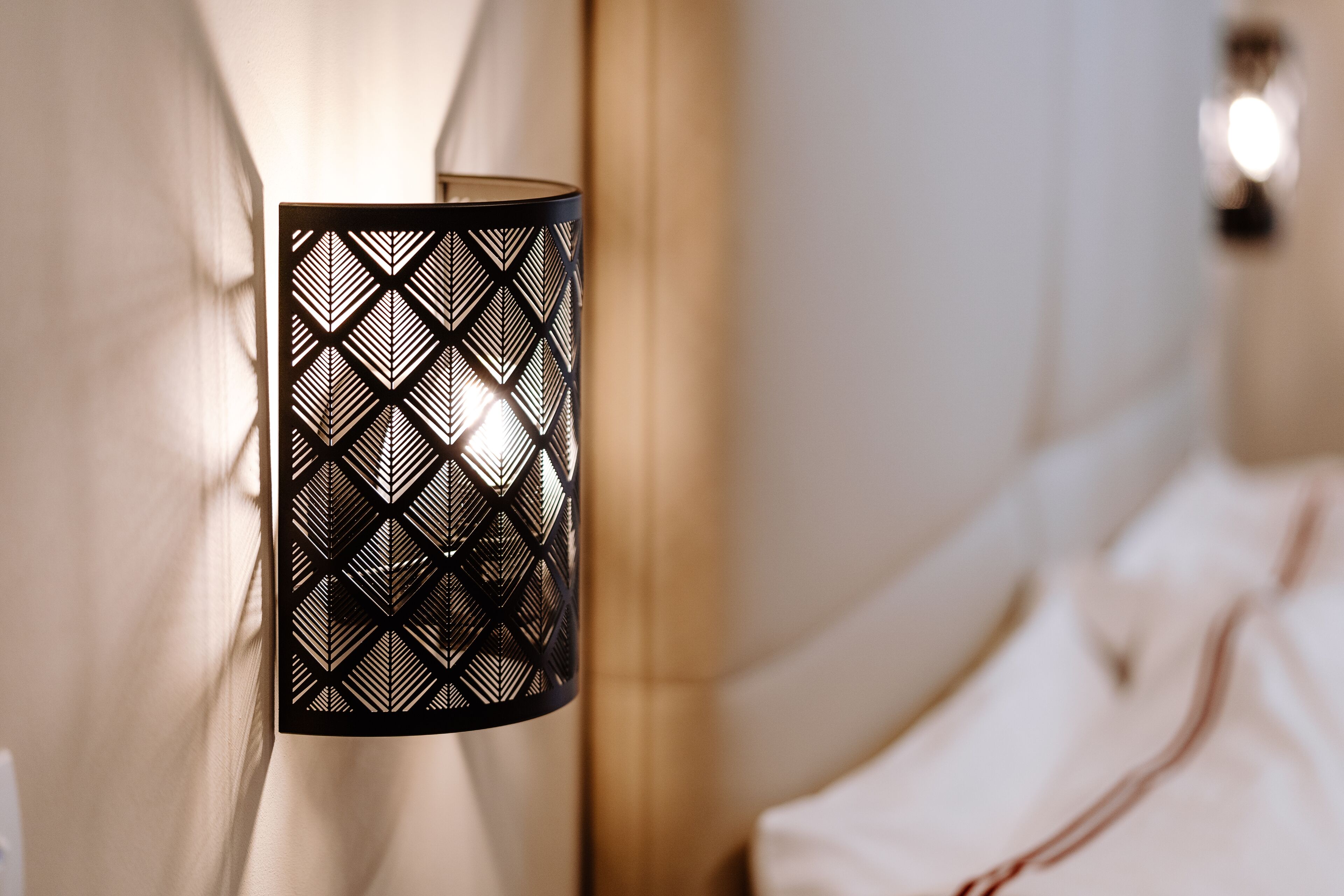 A wall-mounted lamp with a geometric pattern, a bed with white bedding, and a neutral-toned room.
