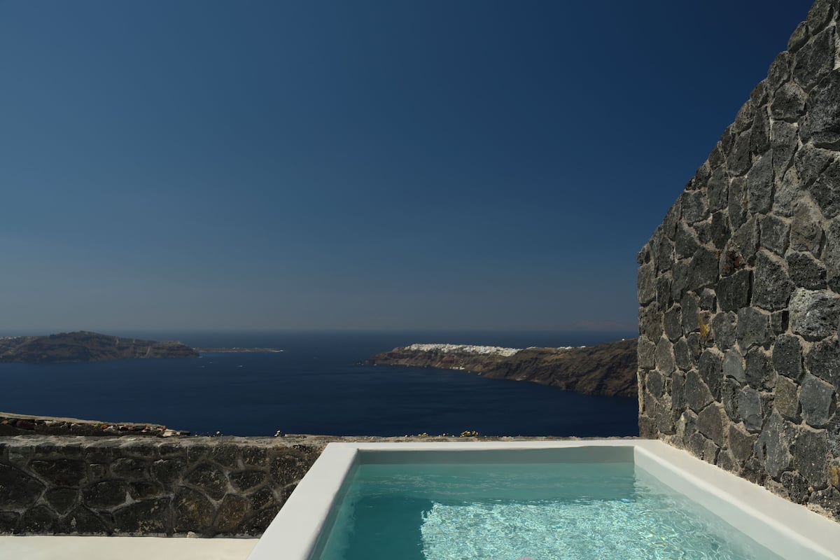 A stone-walled infinity pool with clear blue water, overlooking a rugged coastline and the sea.