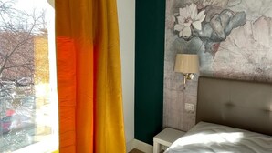 Quarto com cama, mesa de cabeceira, luminária, janela com cortinas e parede com mural floral.