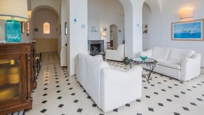 Amplia sala de estar con sofás blancos, una mesa de centro y una chimenea. El espacio cuenta con piso de baldosas y arcos.