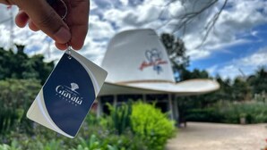A hand holding a keychain with the text "Café Natá" and a white hat with a logo in the background. - Pousada Canto do Gravatá (Penha)