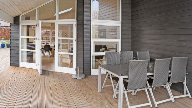 Un patio exterior con piso de madera, una mesa con sillas y un ventanal corredizo que da a un espacio interior.