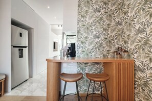 A modern kitchen with a wooden bar and two stools, a refrigerator, and a patterned wall.