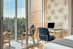 A modern hotel room with a large window, a desk with a laptop, a bed, and a small table with a cup.