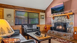 A cozy living room with a stone fireplace, a black leather sofa, and a television mounted on the wall.