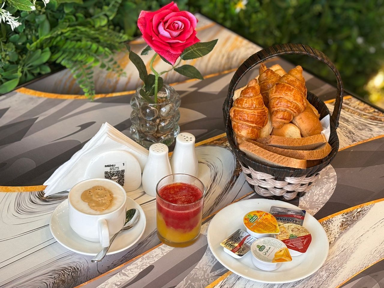 A table with a basket of croissants, a plate of cheese, a cup of coffee, a glass of juice, and a vase with a rose.