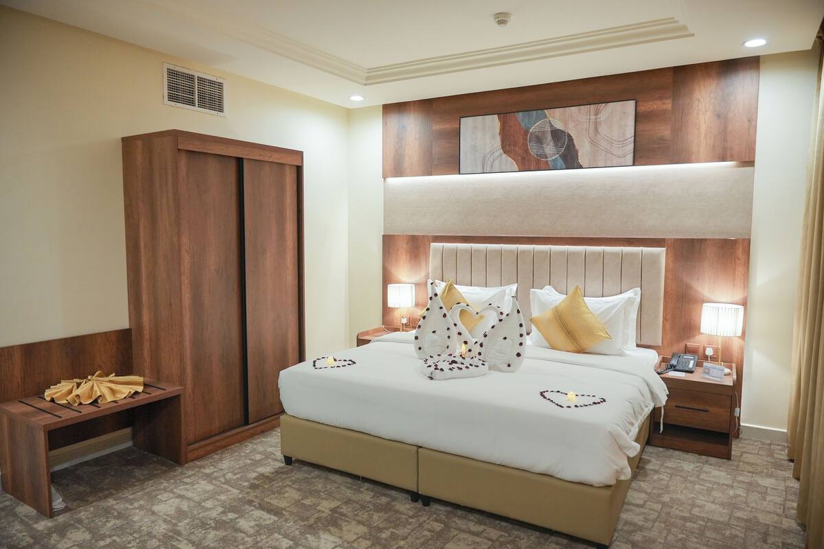A hotel room with a large bed, wooden wardrobe, bedside tables with lamps, and a ceiling vent.