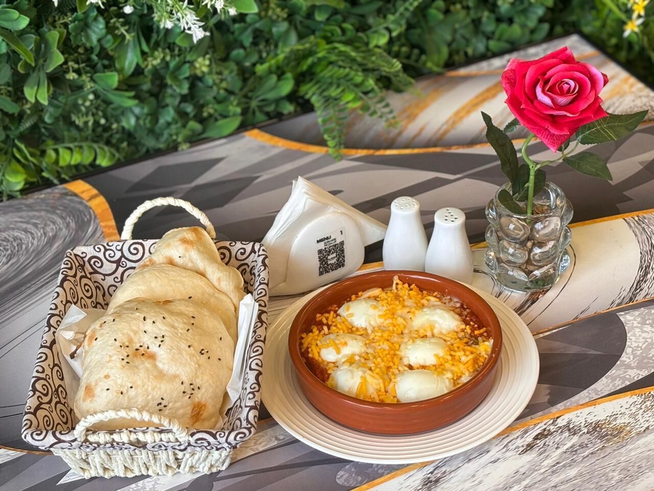 Uma mesa com uma cesta de pão sírio, uma tigela de prato com queijo e ovo, um vaso com uma rosa, saleiro e pimenteiro, e um código QR em um guardanapo.