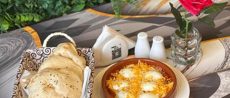 Uma mesa com uma cesta de pão sírio, uma tigela de prato com queijo e ovo, um vaso com uma rosa, saleiro e pimenteiro, e um código QR em um guardanapo.