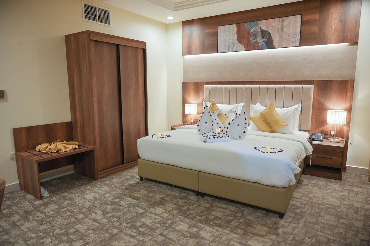 A hotel room with a large bed, wooden wardrobe, bedside tables, and a headboard with artwork.