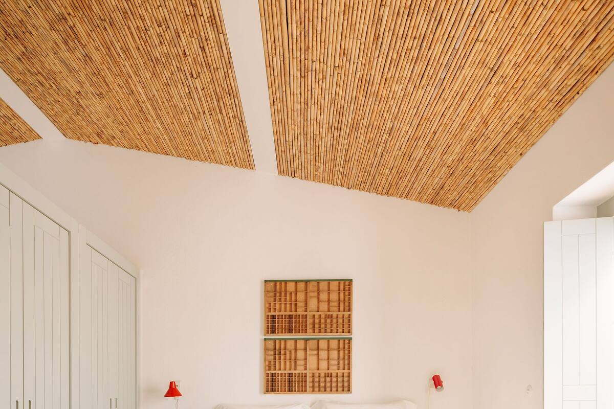 Quarto com cama, um banco de madeira, uma obra de arte na parede e um abajur vermelho.