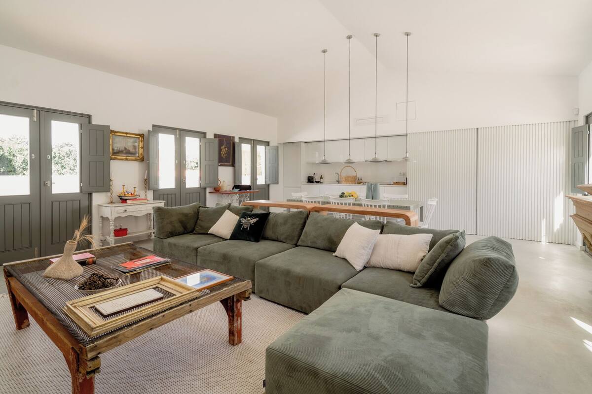 Uma sala de estar moderna com um sofá verde, uma mesa de centro de madeira e uma cozinha ao fundo.