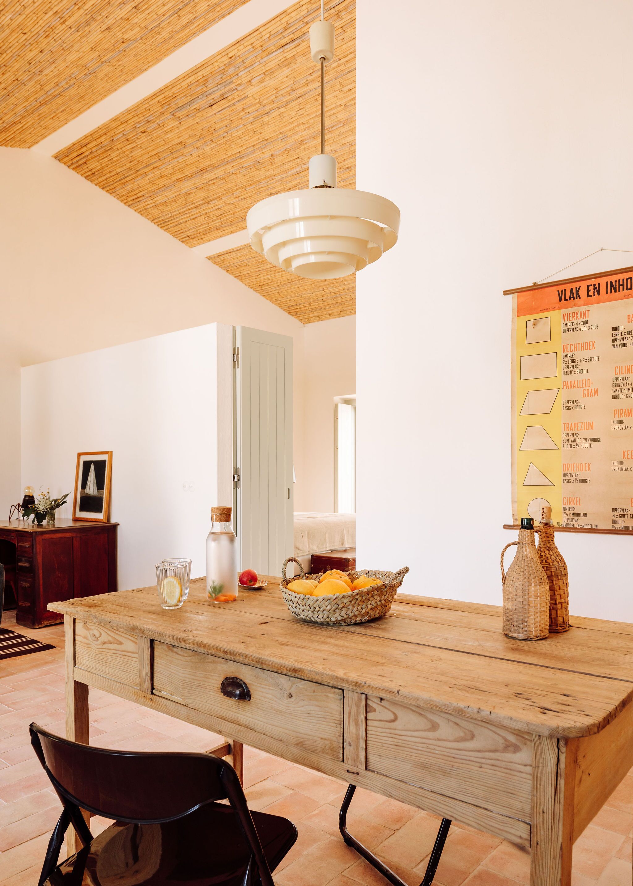 Een houten eettafel met een gevlochten mand vol fruit, een glas water en twee glazen flessen op een houten vloer.