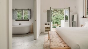 A modern bedroom with a large bed, a wooden bench, a bathtub, and a view of greenery outside. - Casas da Quinta de Cima (Vila Real Santo Antonio)