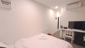 A hotel room with a bed, a desk, a chair, a TV, and an air conditioning unit. - Nomads Homestay Sriracha (Si Racha)