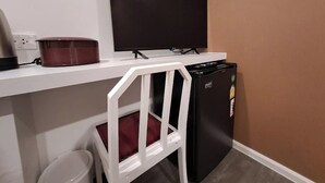 A compact room with a white desk, a black television, a mini fridge, and a white chair. - Nomads Homestay Sriracha (Si Racha)
