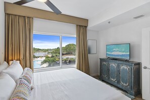 A hotel room with a large bed, a television on a cabinet, and a view of a pool and greenery. - Pool View ~ Wraparound Balcony ~ Tram Included (Miramar Beach)