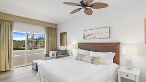 A hotel room with a large bed, a ceiling fan, a window with curtains, a bedside table with a lamp, and a wall-mounted artwork. - Fitness Center ~ Private Beach Access ~ Tram Service Included (Miramar Beach)
