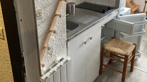 Una cocina compacta con armarios blancos, un taburete de madera y una ventana con vistas a árboles.