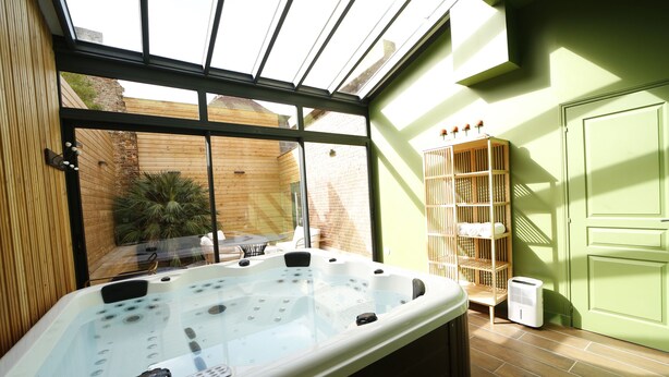 A modern bathroom with a glass-enclosed hot tub, wooden flooring, and a view of a brick wall and greenery outside. - L Arbre d or Appart Hyper Centre SPA Couvert (Cambrai)