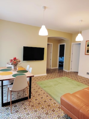 Uma sala de jantar com uma mesa, cadeiras, um vaso com flores, uma televisão de tela plana na parede e um chão estampado.