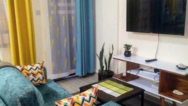 A living room with a sofa, coffee table, TV, and a sliding door with curtains. - Lux Suites Vet Studio Apartments (Ngong)