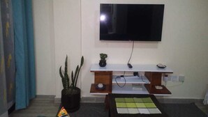 A living room with a flat-screen TV mounted on the wall, a small wooden console table with a plant, and a coffee table with a placemat. - Lux Suites Vet Studio Apartments (Ngong)
