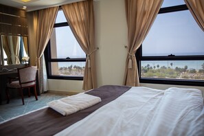 Ein Hotelzimmer mit einem Bett, einem Schreibtisch und einem Stuhl. Große Fenster mit Vorhängen bieten Blick auf den Ozean und Palmen.