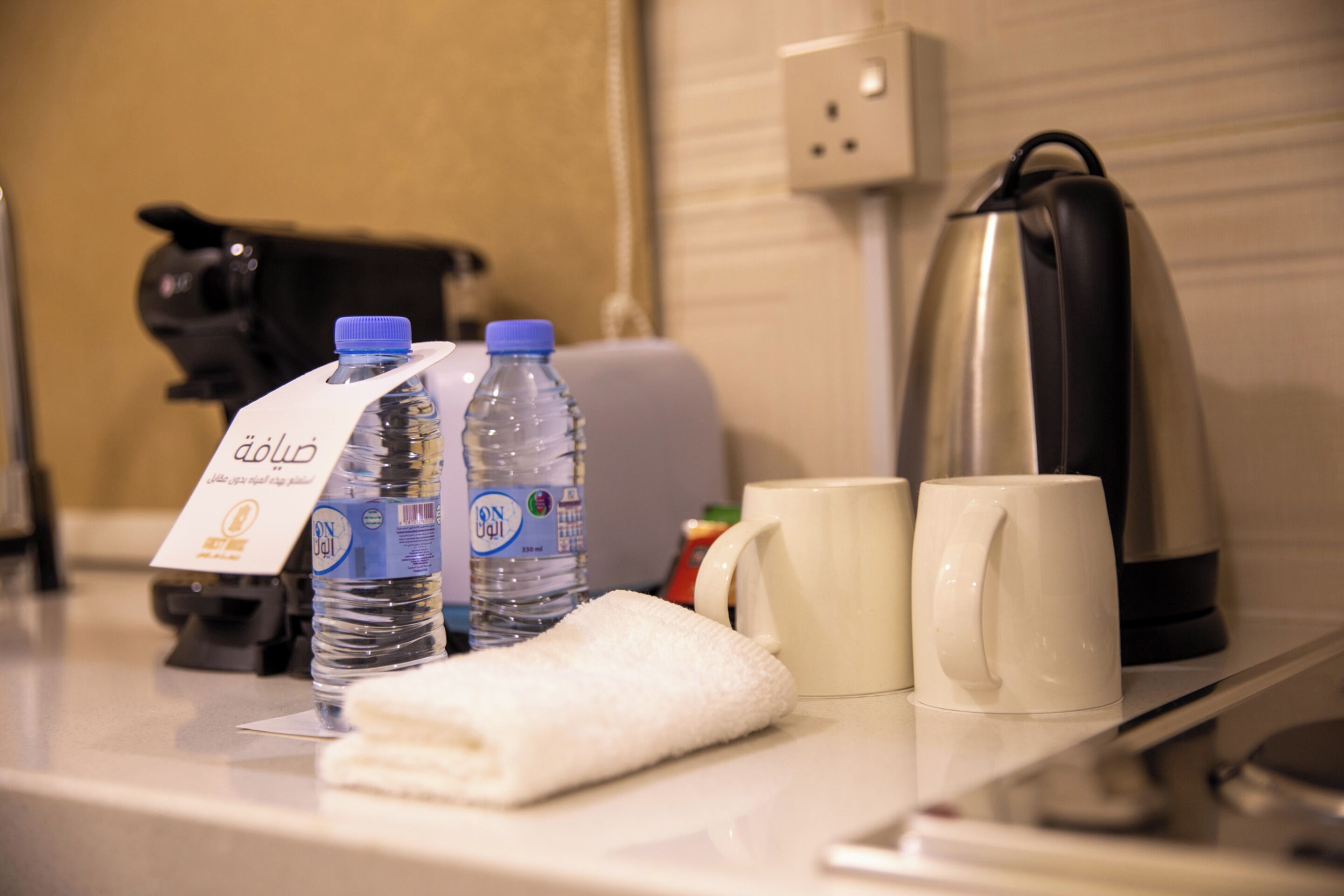 Mostrador de hotel con dos botellas de agua, una toalla, dos tazas blancas, un hervidor y una cafetera.