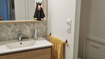 Une salle de bain avec un lavabo blanc, un miroir et des toilettes. Une serviette est accrochée à un porte-serviettes et un récipient bleu se trouve sous le lavabo.