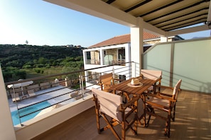A balcony with a pool, wooden furniture, and a view of a hillside. - Michelangelo Suites and Apartments (Parga)