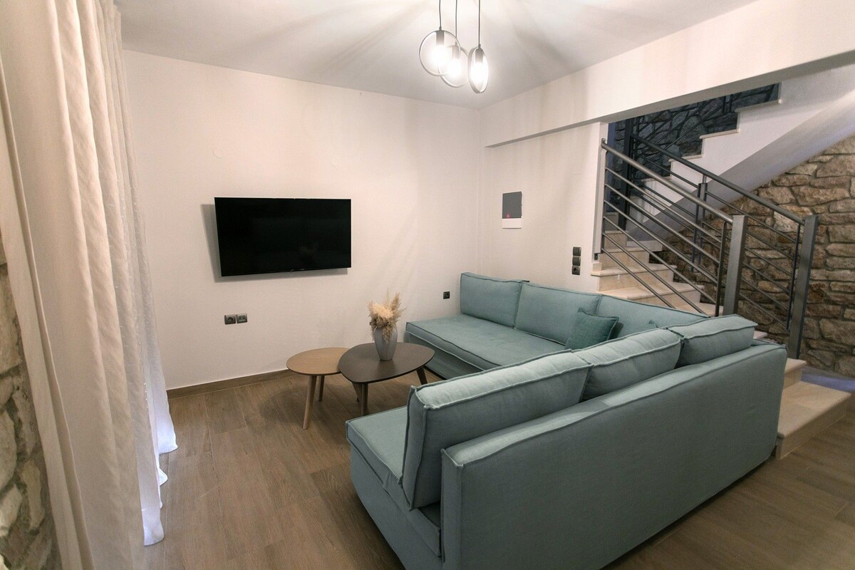 A modern living room with a large L-shaped sofa, a flat-screen TV mounted on the wall, a small round table, and a staircase with a stone wall.