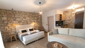 A bedroom with a stone wall, a bed with white linens, a wooden bench, a white sofa, and a small table with a hat. - Michelangelo Suites and Apartments (Parga)